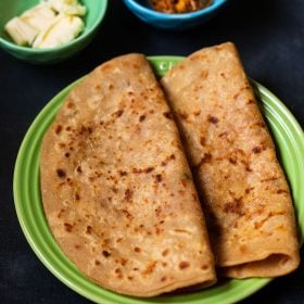 aloo paratha folded and placed on a green ceramic plate on a dark bluish board. sides of butter cubes and mango pickle served in two small ceramic bowls.