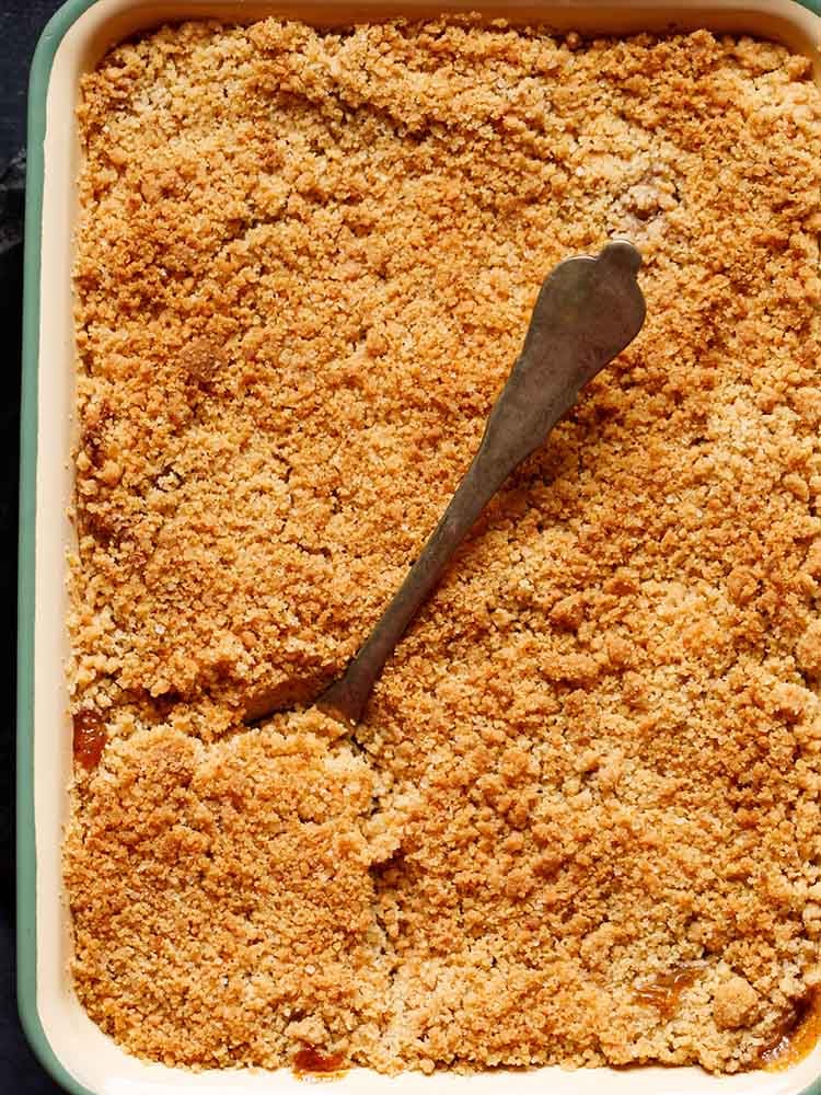 top shot of apple crumble in the baking tray with a brass serving spoon inside on lower left hand side of the crumble