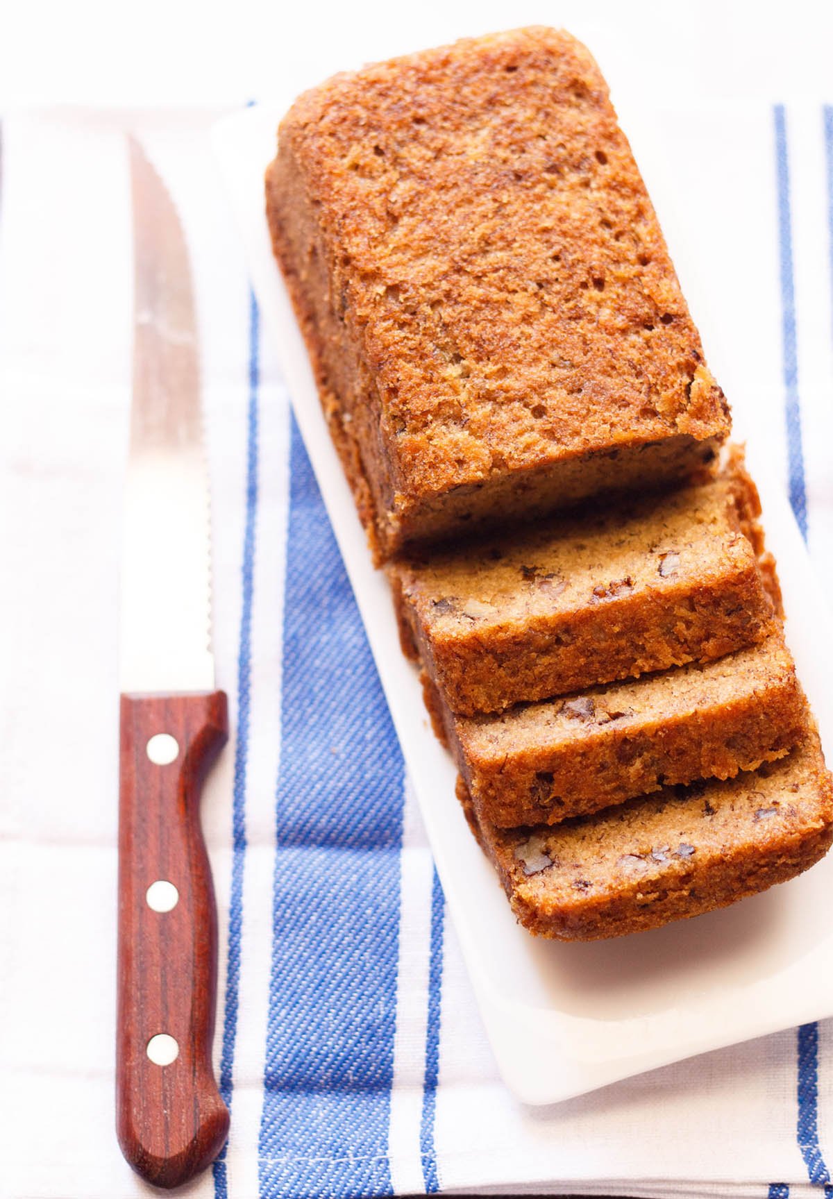 loaf of sliced eggless banana cake on white tray with serrated wooden knife on side