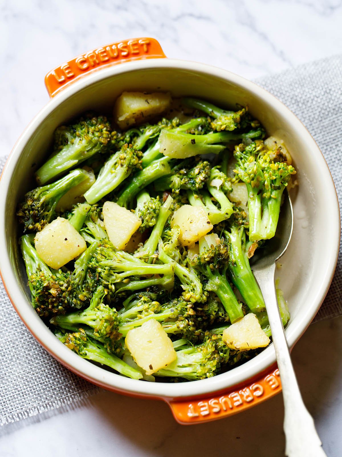 sauteed broccoli in a le creuset cream ceramic pan with orange handles with a spoon inside placed on a folded light gray jute fabric