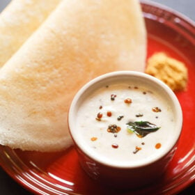 a small white bowl of coconut chutney on a red plate with two dosa