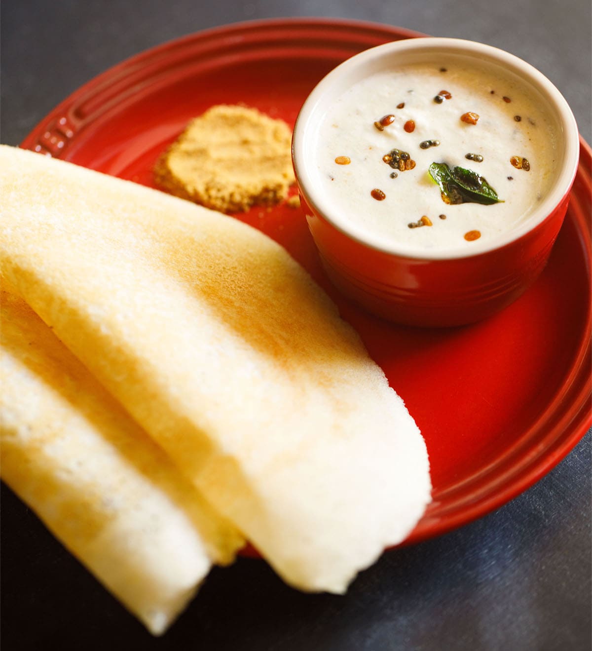 small white bowl of coconut chutney on a red plate with two dosas