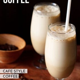 side shot of cold coffee in two glass with a white straw in one glass with coffee beans in a dark orange bowl and some coffee beans on a wooden board with a text layover