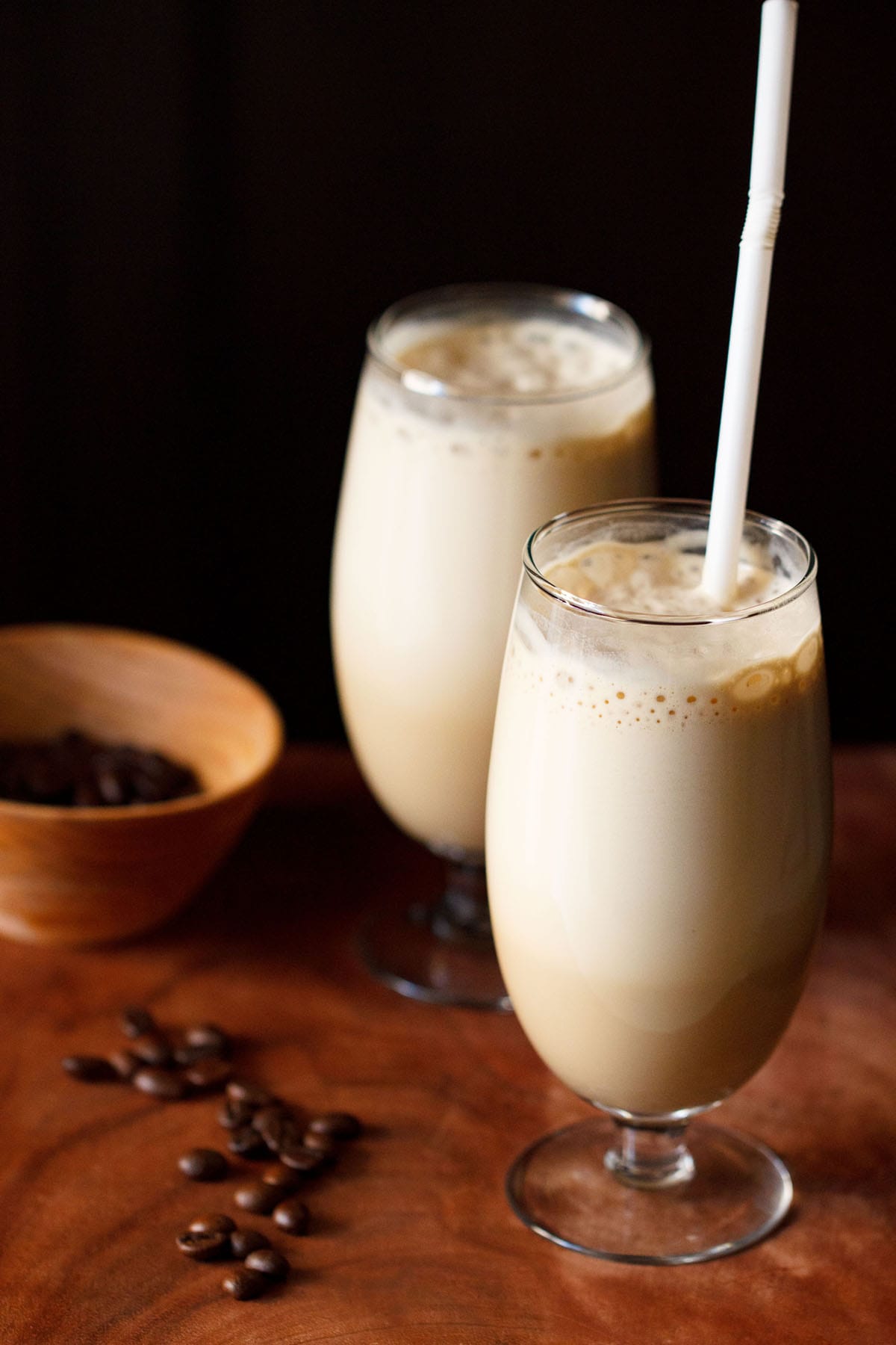 side shot of cold coffee in two glass with a white straw in one glass with coffee beans in a dark orange bowl and some coffee beans on a wooden board