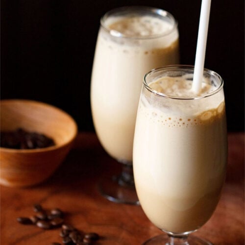 side shot of cold coffee in two glasses with a white straw in one glass with coffee beans in a dark orange bowl and some coffee beans on a wooden board