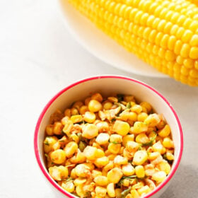 corn salsa served in a dark pink rimmed white bowl on a white background with a steamed corn cob at the side