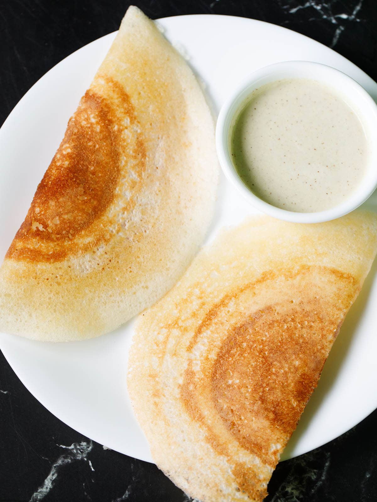 two crisp and golden dosa with a bowl of coconut chutney on a white plate