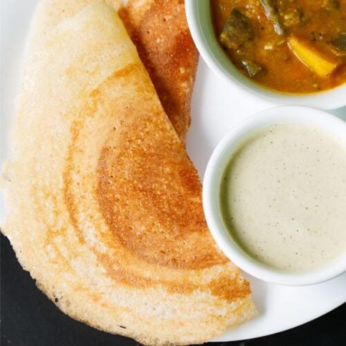 dosa on white plate next to white bowls filled with sambar and coconut chutney