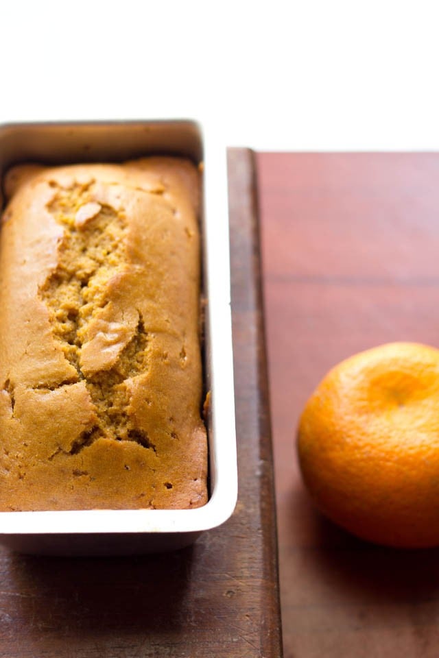 eggless orange cake in a loaf pan with a orange next to it on a brown table