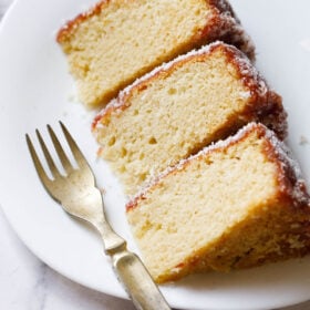 close up shot of thick slices of eggless vanilla cake on a square shaped white plate with a brass fork on the plate.