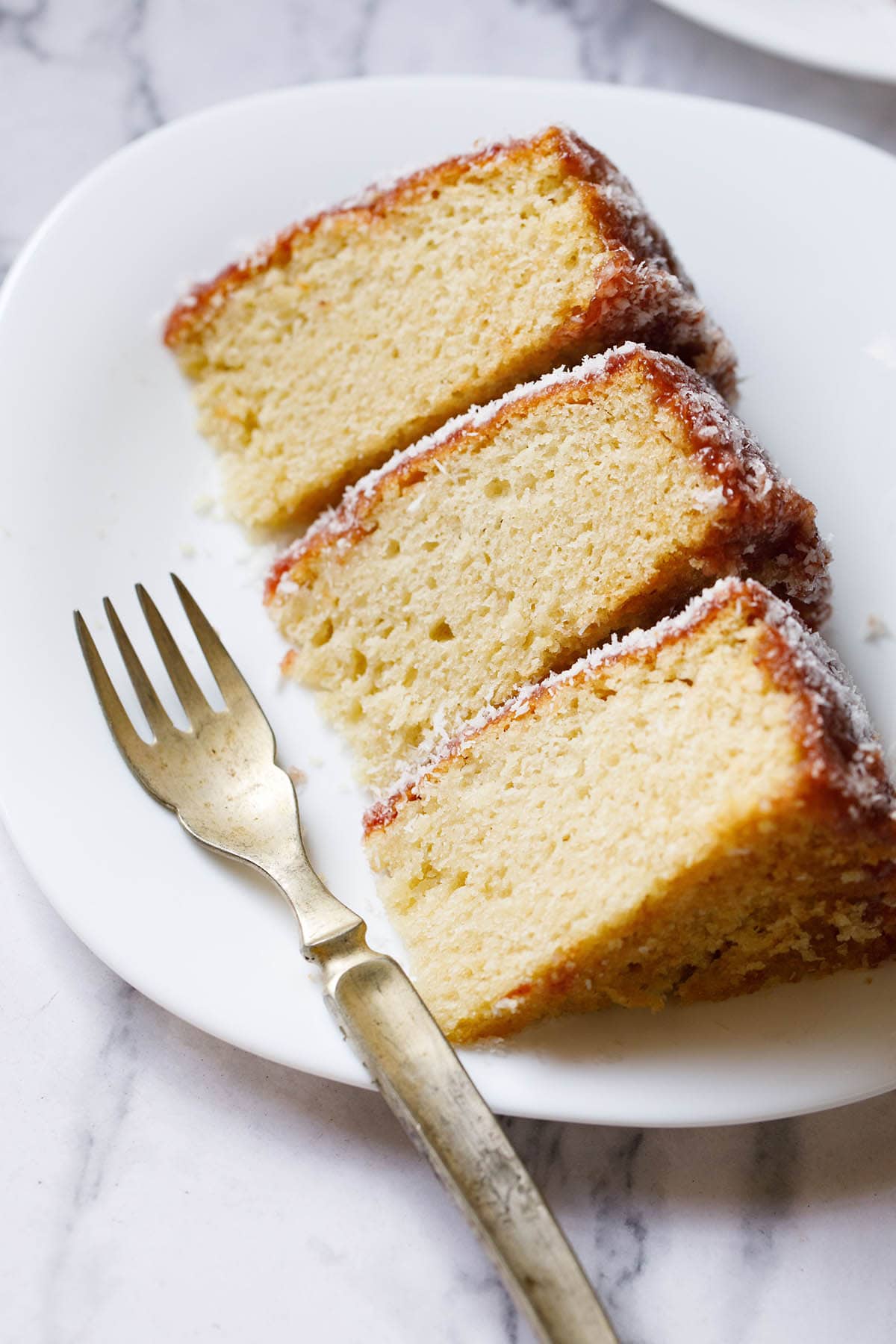 close up shot of thick slices of eggless vanilla cake on a square shaped white plate with a brass fork on the plate.