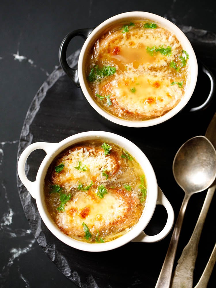 french onion soup in two cream colored soup bowls with spoons on a black slate tray. soup garnished with some parsley.