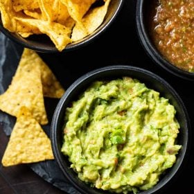guacamole served in a black wooden bowl with a side black bowl of nachos and tomato salsa