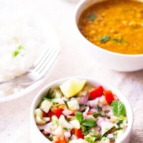 kachumber salad in a small white bowl on a table next to plate of rice and a bowl of lentils
