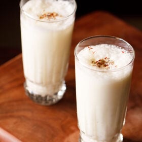 sweet lassi topped with crushed cardamom powder and saffron in two tall glasses on a wooden board