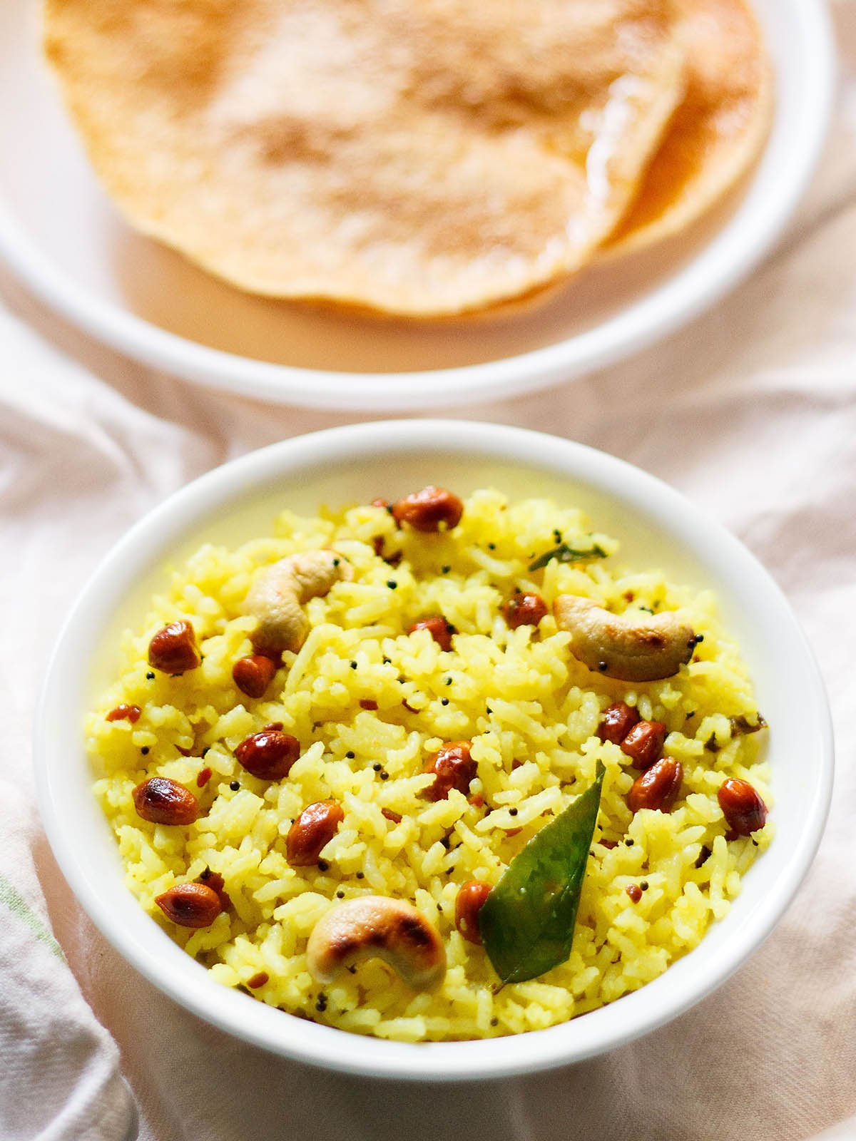 bright yellow colored lemon rice or chitranna served in a white bowl on a white cotton cloth with a side of fried papaddums