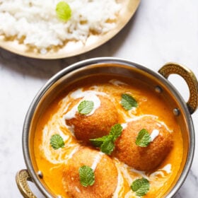 top shot of 3 malai kofta with mint leaves on top in a silver serving dish with handles on a white marble
