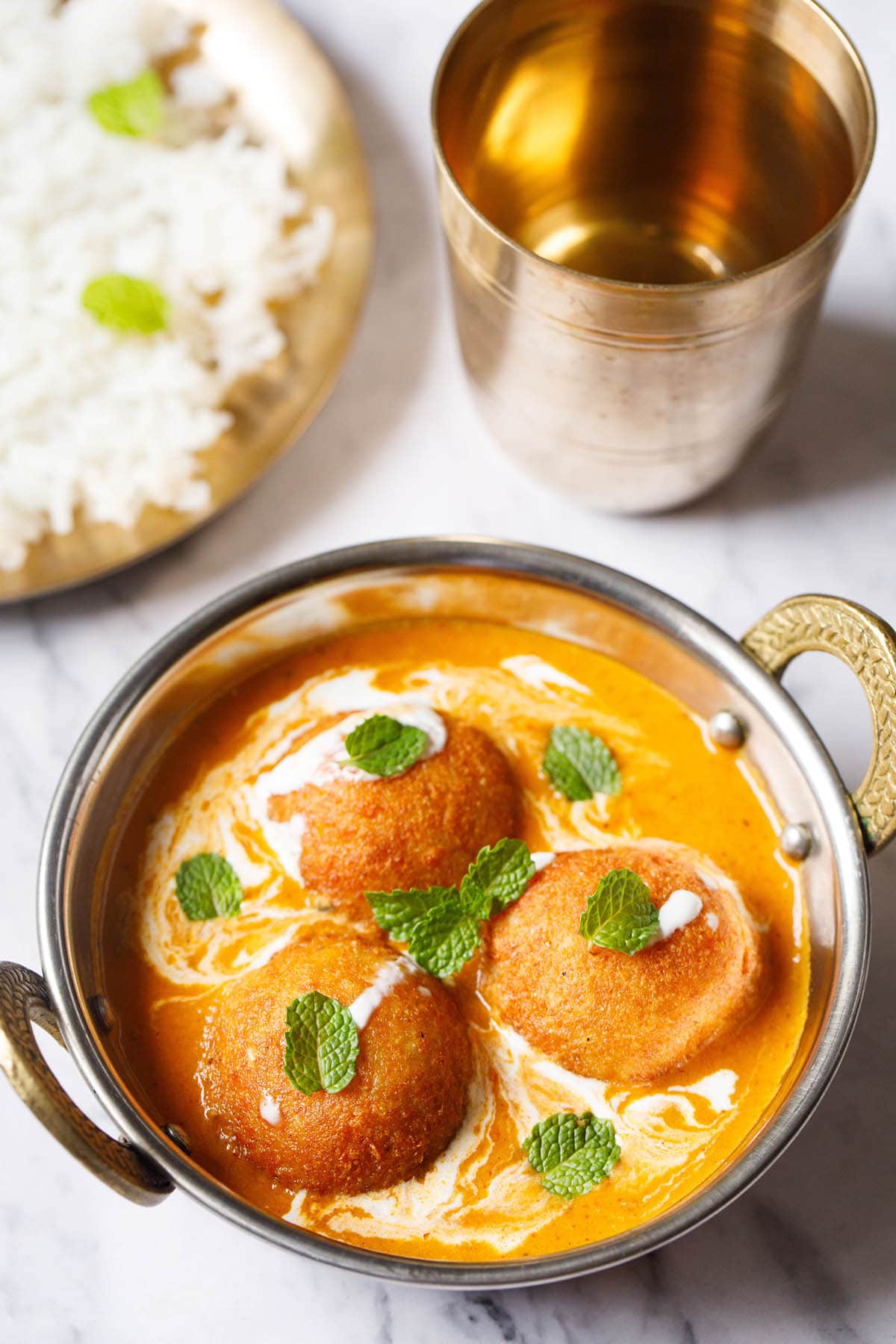3 malai kofta garnished with mint leaves in a silver serving dish with handles on a white marble