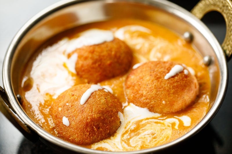 three malai kofta placed on the gravy with a drizzle of cream on top