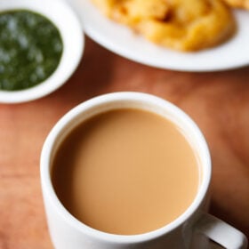 masala chai filled in a white cup with a side of onion pakora in white plate and green chutney in small white bowl