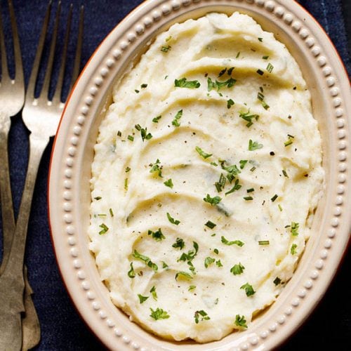 mashed potatoes served in an oval ceramic bowl kept on a slate grey wooden board. garnished with some finely chopped parsley and rosemary.