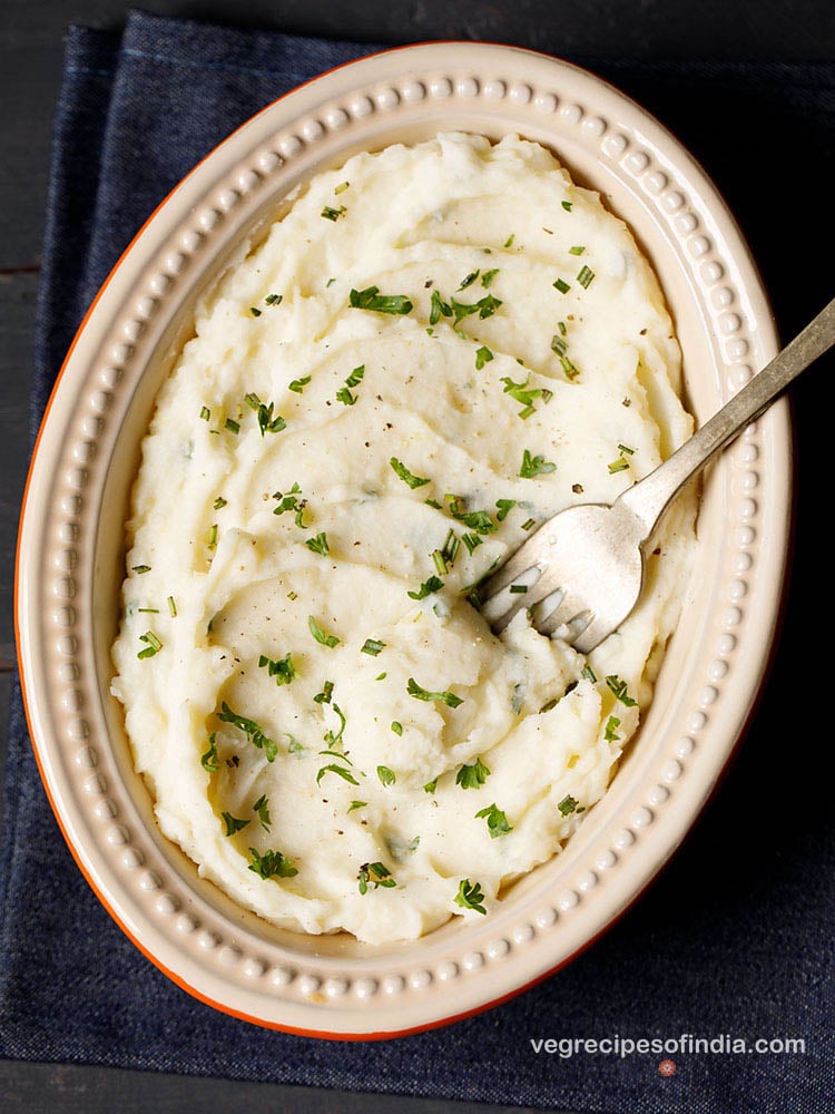 mashed potatoes served in an oval ceramic bowl with a fork in it. bowl is kept on a slate grey wooden board. garnished with some finely chopped parsley and rosemary.