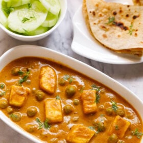 Top shot of matar paneer on white rectangle plate next to sliced cucumber in a white bowl and folded two roti in a white plate sitting on white marble table
