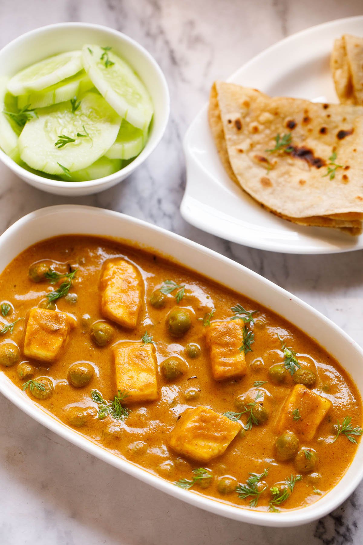 Top shot of matar paneer on white rectangle plate next to sliced cucumber in a white bowl and folded two roti in a white plate sitting on white marble table