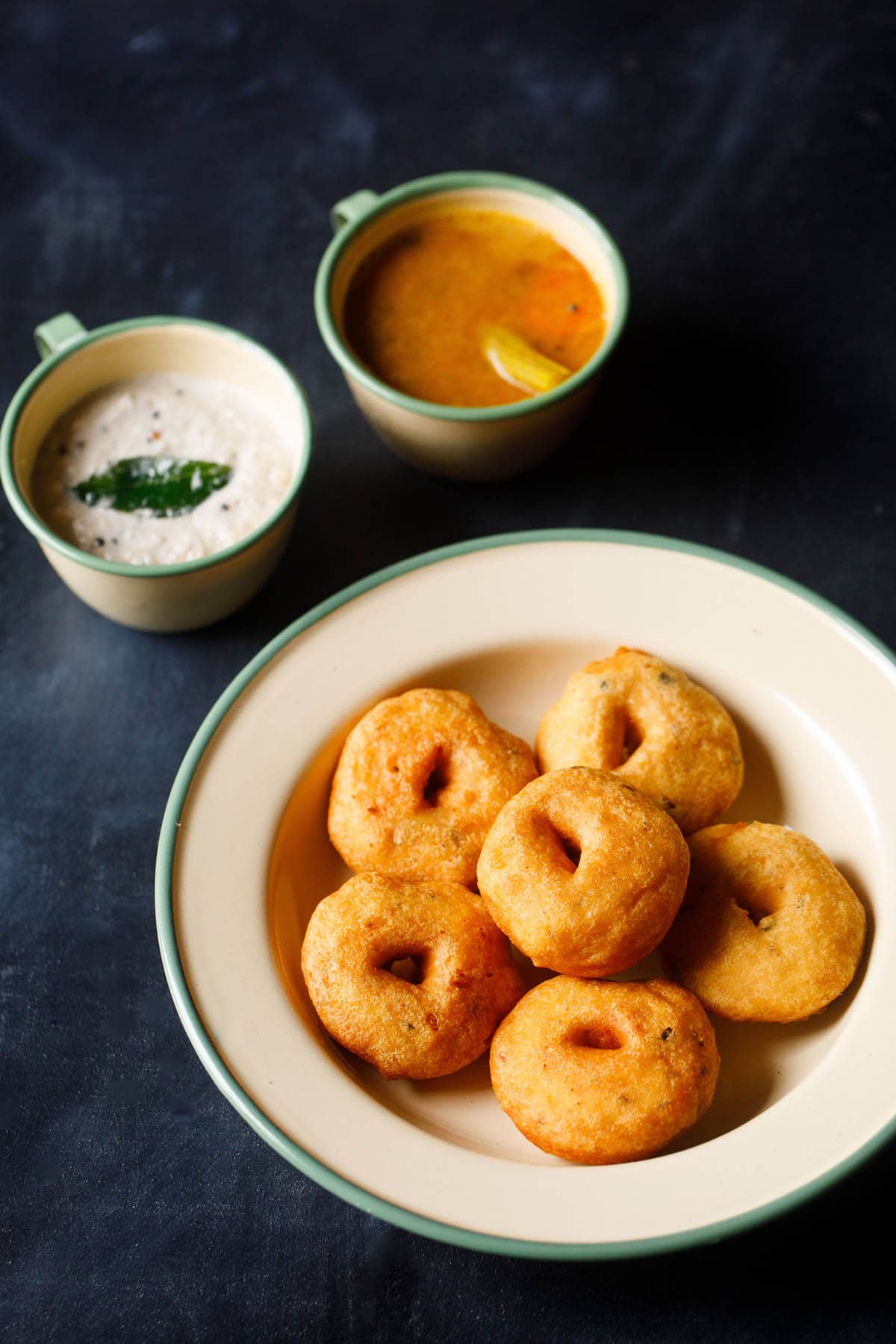 top shot of medu vada stacked neatly in green lined beige colored plate with cups filled with coconut chutney and sambar on the top