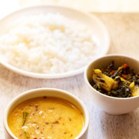 moong dal recipe in white bowl with a plate of steamed rice and a vegetable dish in the background