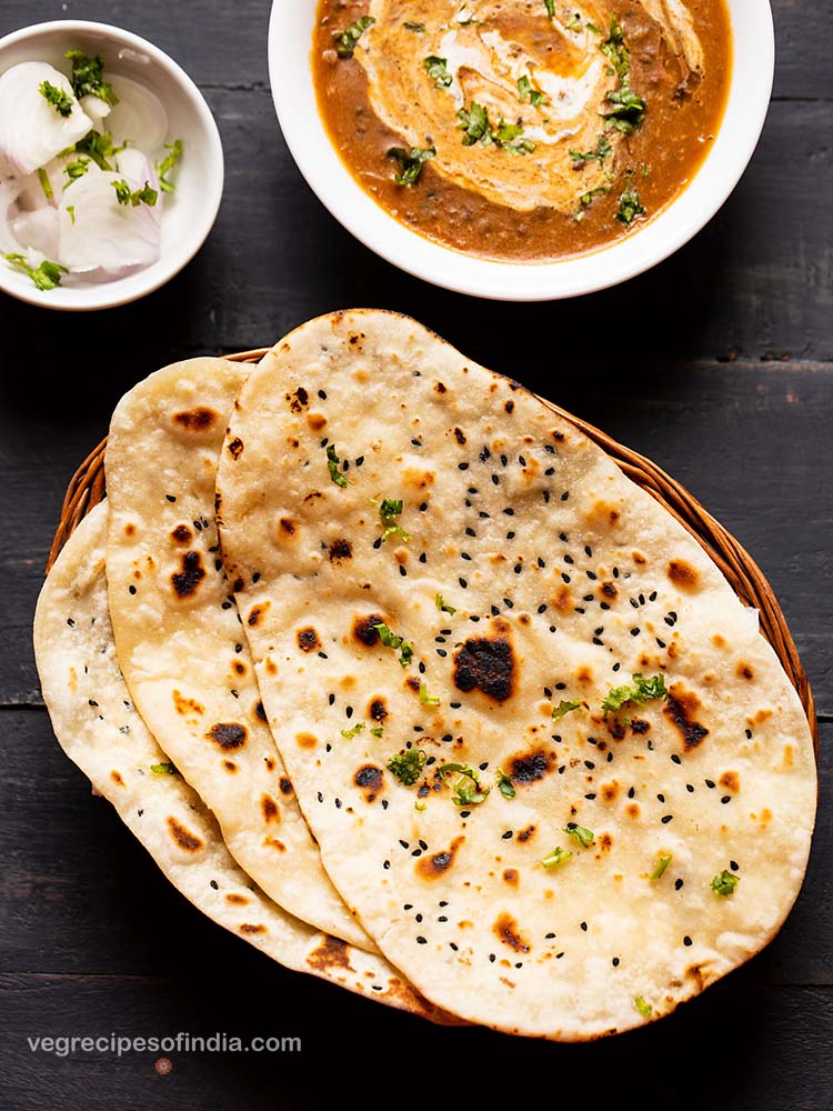 naan kept on top of each other in a cane basket. served with dal makhani in a white bowl and side of sliced onions garnished with coriander leaves in a small bowl.