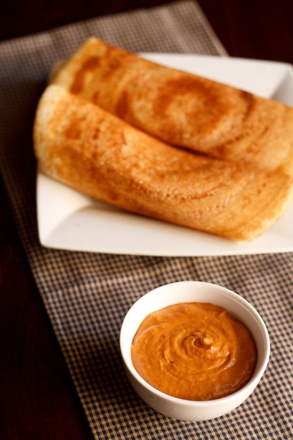 onion tomato chutney served in a small white bowl with a platter of dosas kept on the top side.
