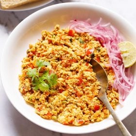 Paneer Bhurji served in a shallow white bowl garnished with one coriander sprig with a spoon inside the bhurji and topped with some sliced onions and a wedge of lemon