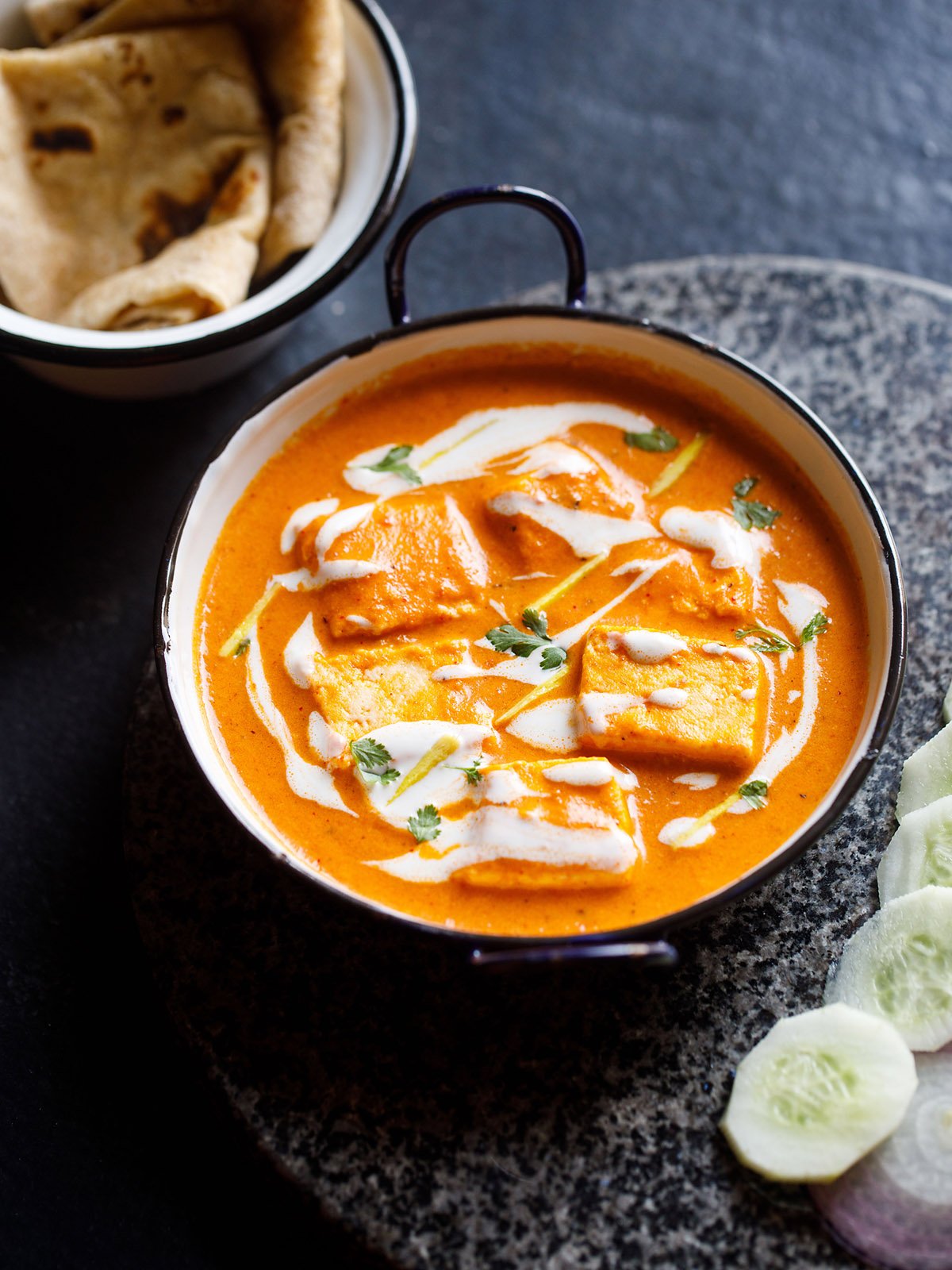 paneer butter masala served in a blue rimmed white pan, garnished with cream and cilantro with a side of sliced cucumber