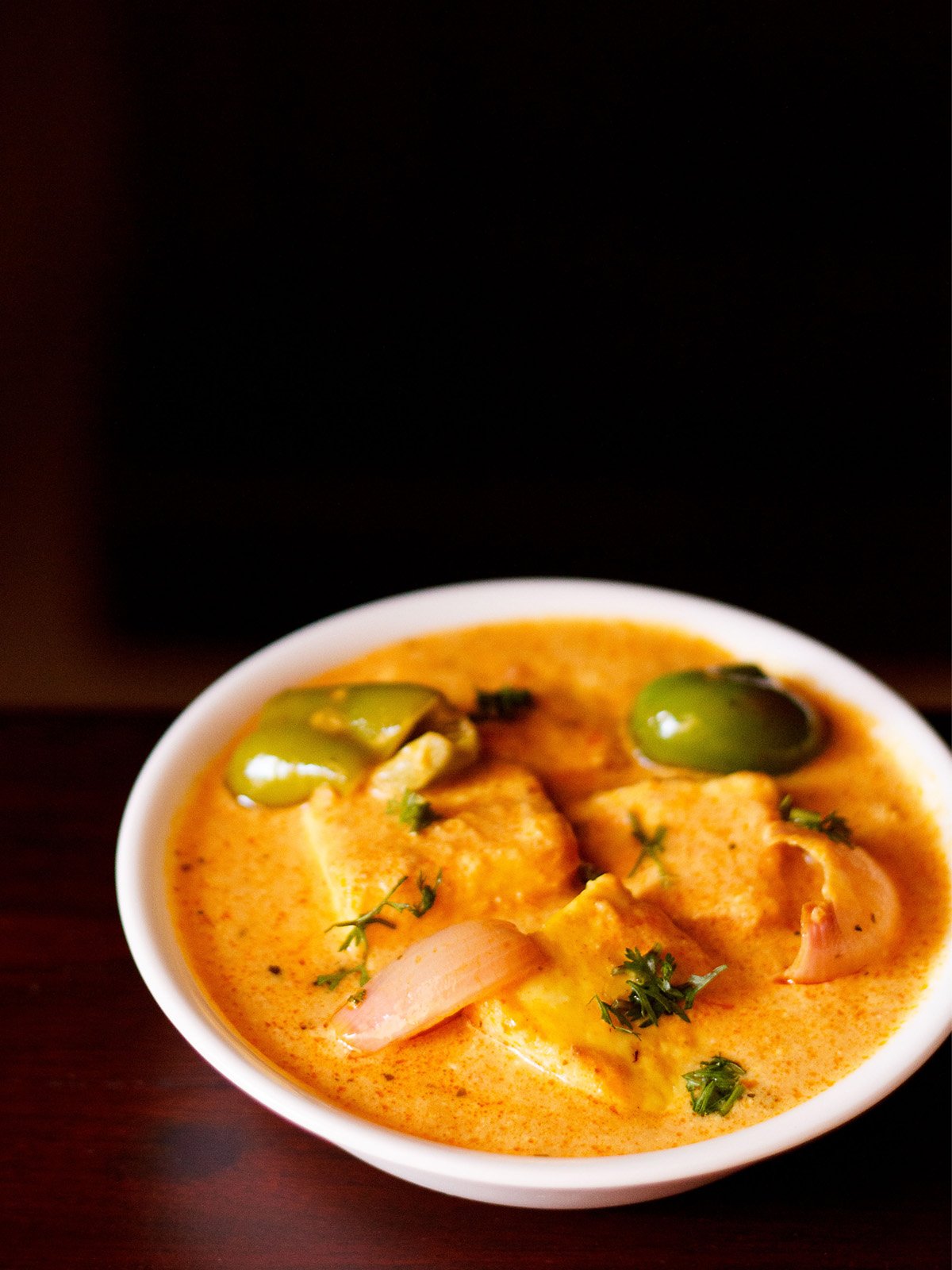 paneer tikka masala served in a white bowl garnished with some coriander leaves on a dark mahogany table.