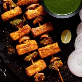 wo grilled paneer tikka skewers kept on a black slate board with a bowl of cilantro dip on top right, one lemon wedge below the bowl and a few onion rounds near the lemon wedge