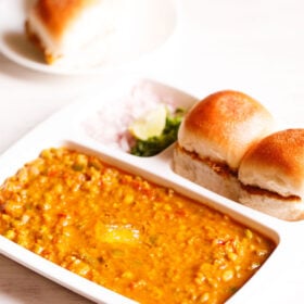 pav bhaji served in a rectangular serving tray with buttered pav and chopped onions, cilantro and lemon wedges on a white table