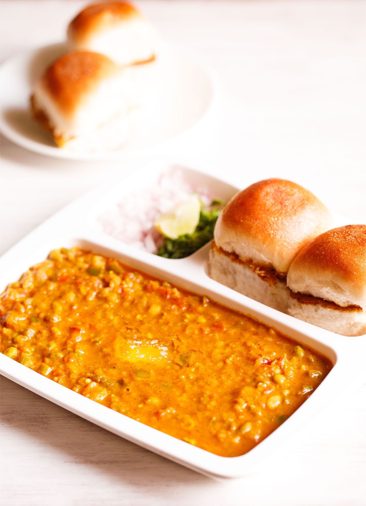 pav bhaji served in a rectangular serving tray with buttered pav and chopped onions, cilantro and lemon wedges on a white table