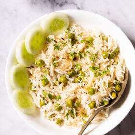 overhead shot of green peas pulao in a white shallow bowl with a brass spoon and cucumber slices placed at the left side on a white marble table