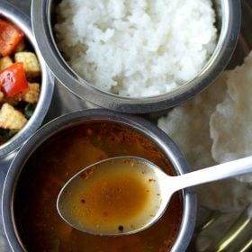 milagu rasam served in a steel bowl with some in a steel spoon on a plate with a bowl of vegetable stir fry, steamed rice and text layover.
