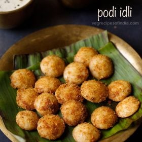 podi idli served on a banana leaf placed on a brass plate with a brass bowl of coconut chutney and brass bowl of sambar kept on the top left side and text layover.