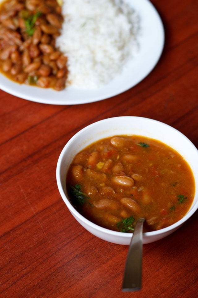 rajma masala in a white bowl with a spoon and a plate filled with rice and rajma curry placed on top