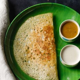 rava dosa served in a green plate with a side of two bowls having sambar and coconut chutney on a dark black-blue board
