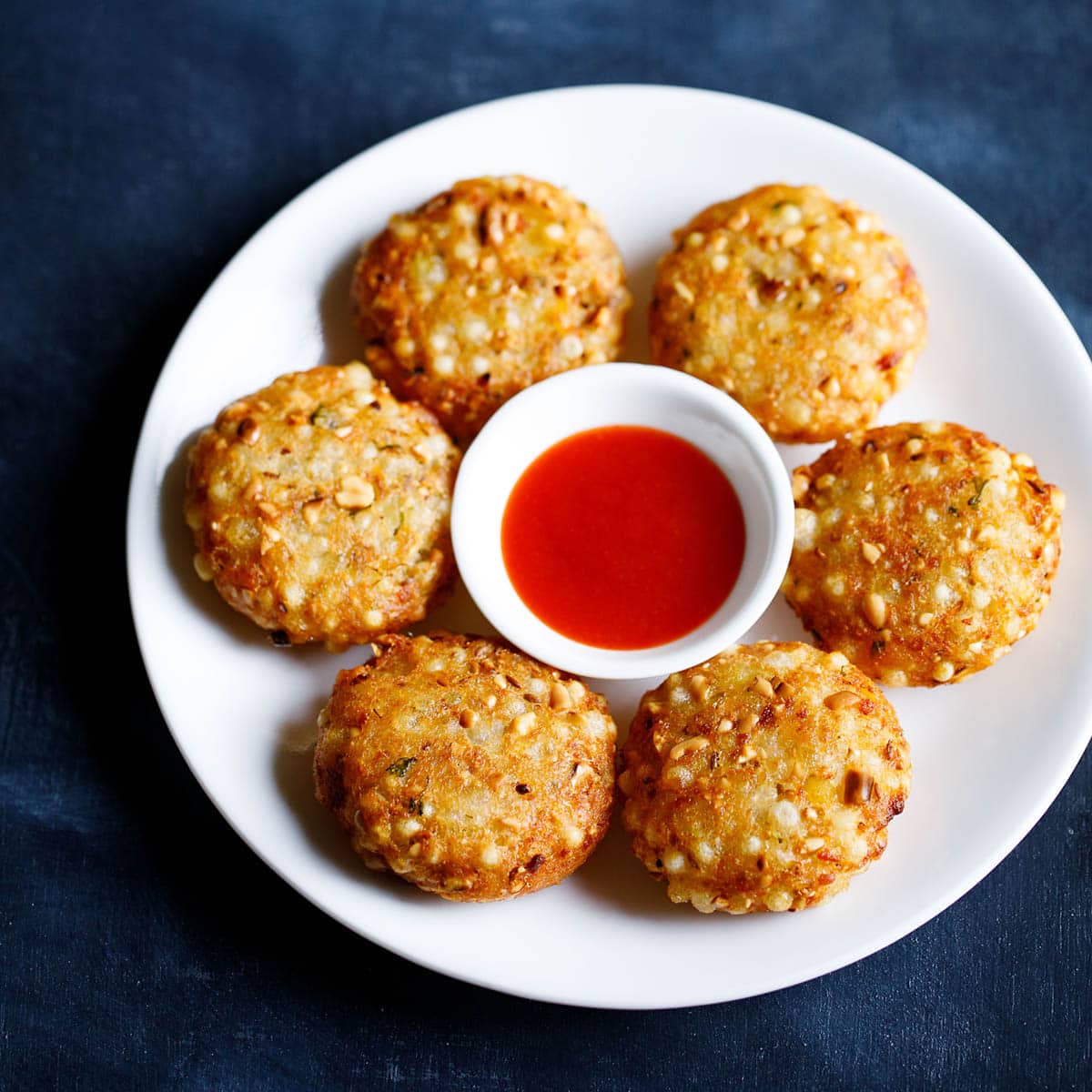 overhead shot of sabudana vada arranged in a hexagon shape with a a red colored tamarind chilli chutney in a center bowl on a white plate on a dark blue board