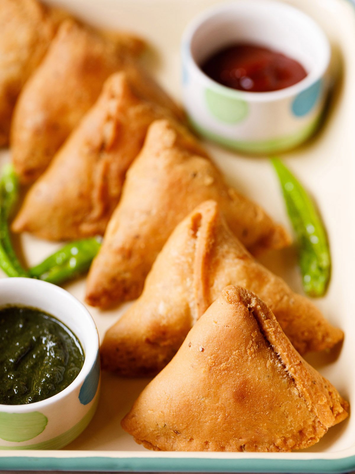 samosa stacked neatly on a cream tray with small bowls of chutney and salted fried green chillies