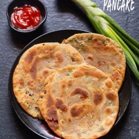 scallion pancakes placed on a black ceramic plate on a black board with sriracha sauce served in a small black bowl on left side and a stalk of spring onion placed on the right side of the plate.