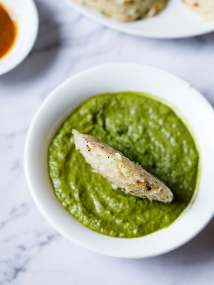 rava Idli dipped in a white bowl having coriander chutney placed on a white marble backdrop