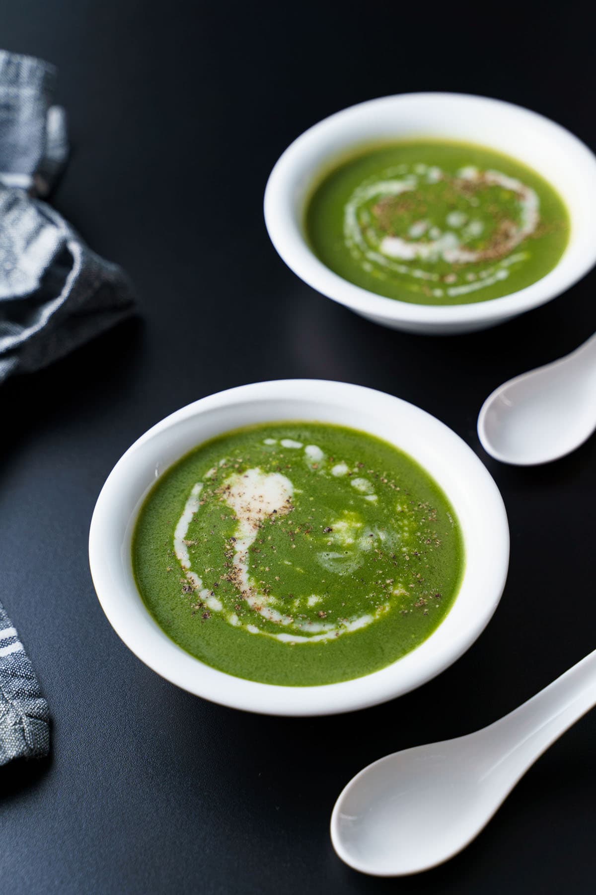spinach soup drizzled with coconut cream on top in two white bowls and two white spoons placed next to bowls on a black board