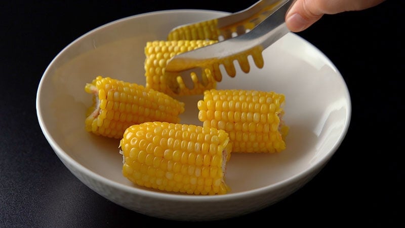 sweet corn cobs being placed with pasta tongs in a white bowl
