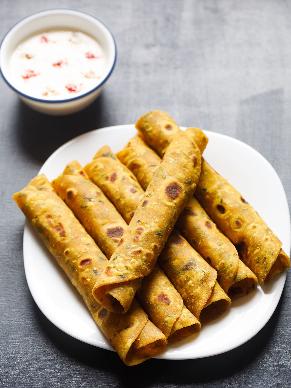 a thepla roll placed in a slant direction over five stacked methi thepla rolls on white plate on blue black board with a side of raita on top left